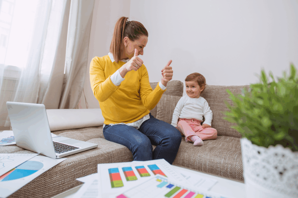 Mutter arbeitet im Homeoffice und zeigt Daumen hoch, während ihr kleines Kind neben ihr auf dem Sofa sitzt – Symbol für Vereinbarkeit von Familie und Beruf.