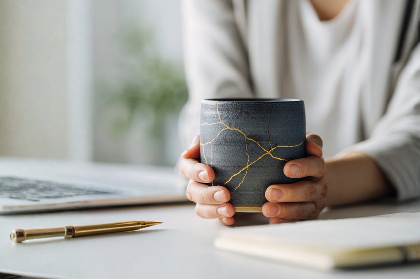 Kintsugi-Tasse mit goldenen Rissen in den Händen einer Frau am Schreibtisch – Symbol für berufliche Brüche im Berufsleben. 
