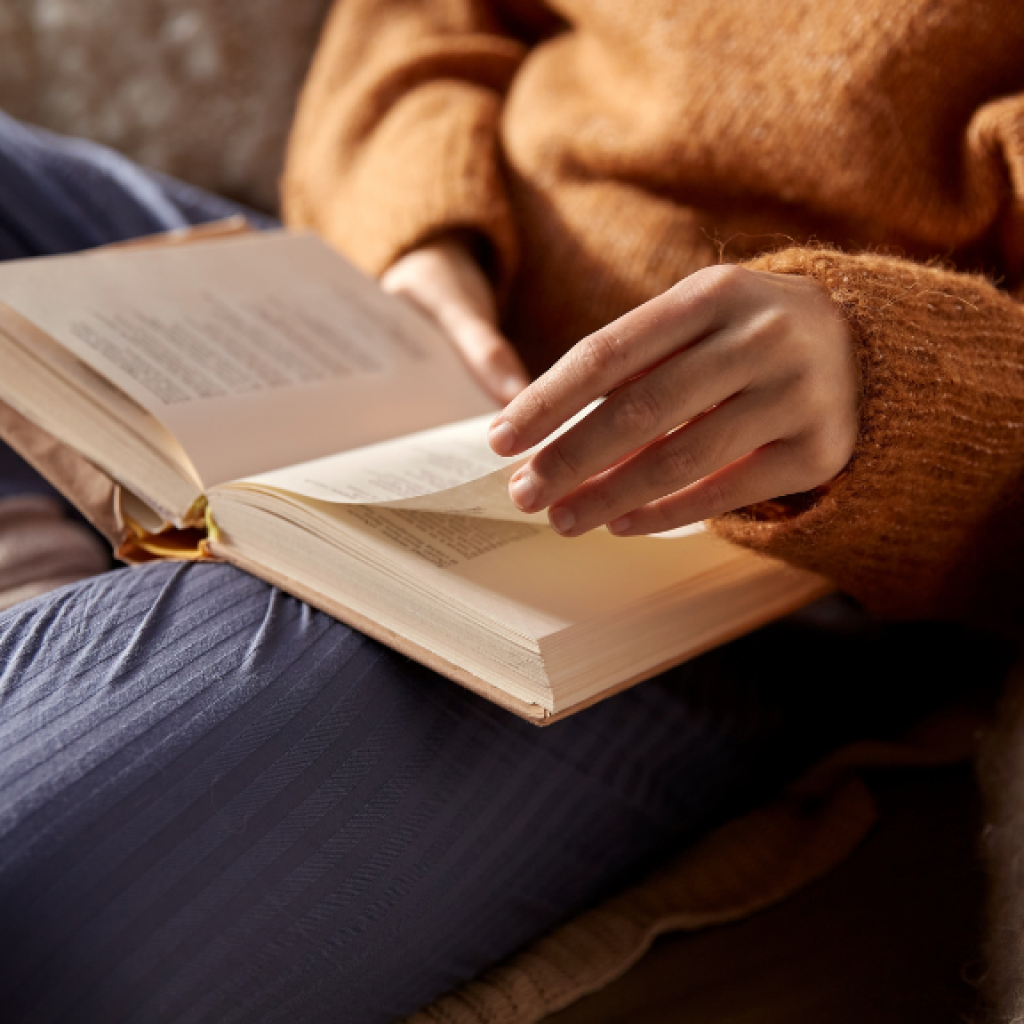 Eine Frau sitzt gemütlich auf dem Sofa und blättert in einem offenen Buch.