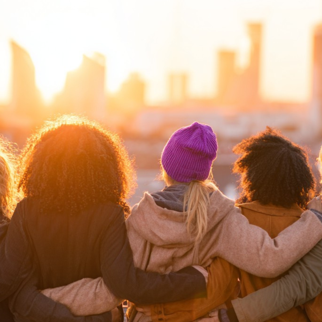 Weltfrauentag 2026: Fünf Frauen verschiedener Herkunft stehen Arm in Arm auf einem Dach und blicken bei Sonnenuntergang gemeinsam auf eine moderne Skyline; eine Frau trägt eine violette Mütze als Symbol für die Frauenbewegung.
