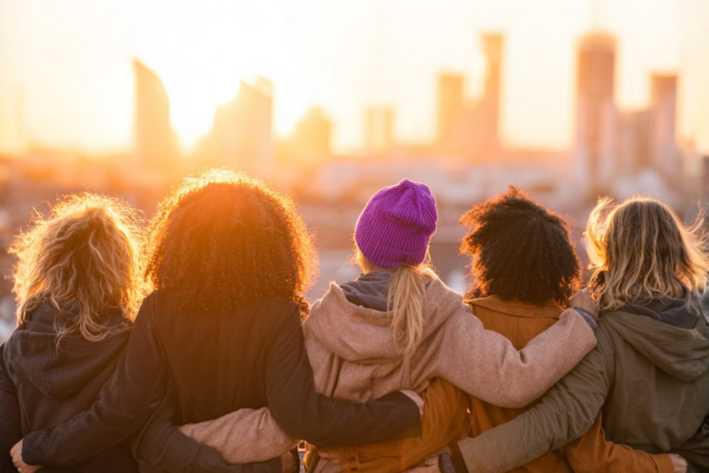 Weltfrauentag 2026: Fünf Frauen verschiedener Herkunft stehen Arm in Arm auf einem Dach und blicken bei Sonnenuntergang gemeinsam auf eine moderne Skyline; eine Frau trägt eine violette Mütze als Symbol für die Frauenbewegung.